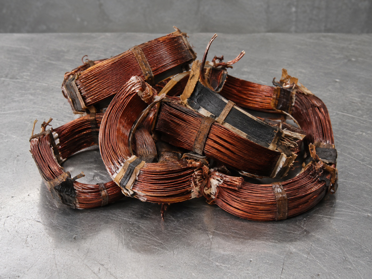 Illustrative example: A small heap of extracted copper electric motor windings on a steel workbench, bundled curved stiff varnished reddish copper wire loops, some black insulation remnants, realistic motor winding copper, not shiny new cable or copper granules.